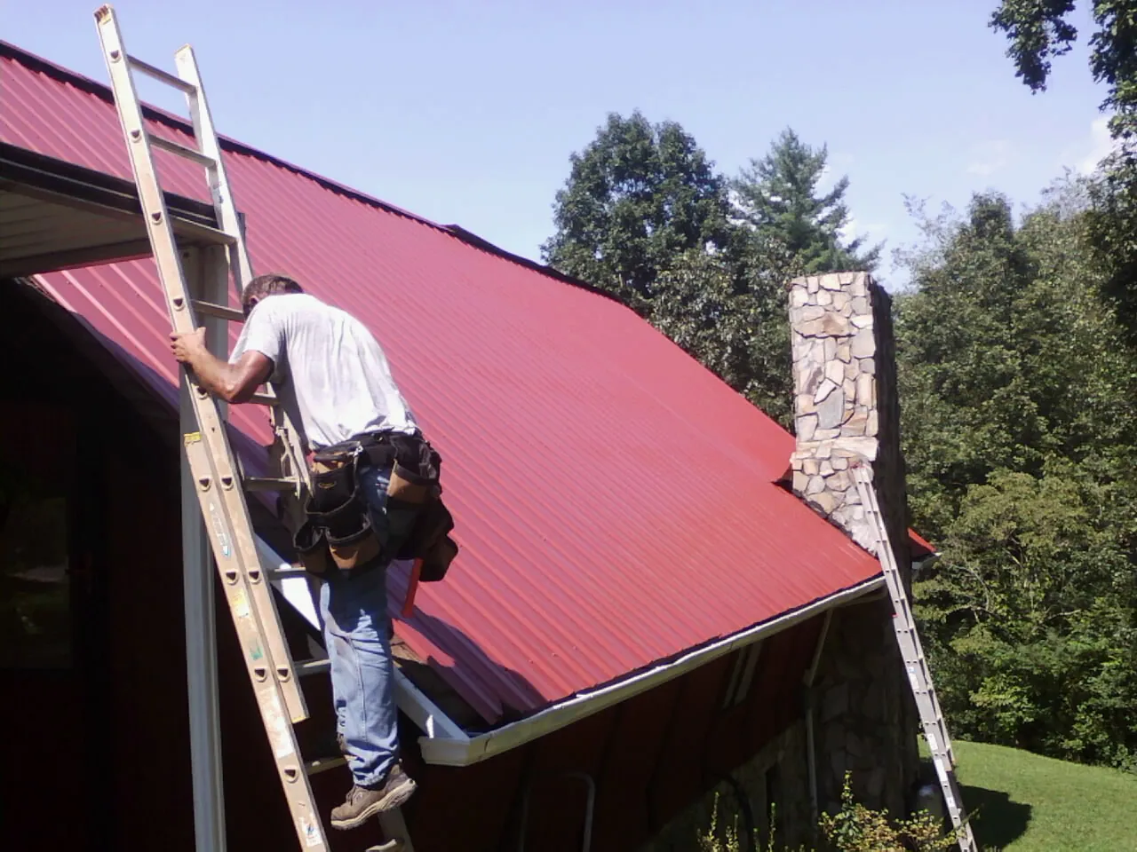 Roofer on a ladder beside a red metal roof and stone chimney — Soffit & Fascia Repair by Roofing Brothers in Issaquah